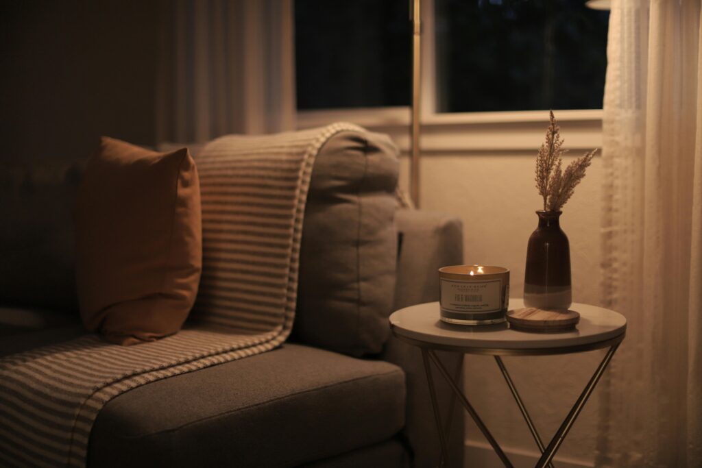 Przytulny kącik do czytania w stylu hygge z beżowym fotelem, ciepłym oświetleniem świecy na okrągłym stoliku, suszonymi trawami w ciemnym wazonie przy oknie wieczorem