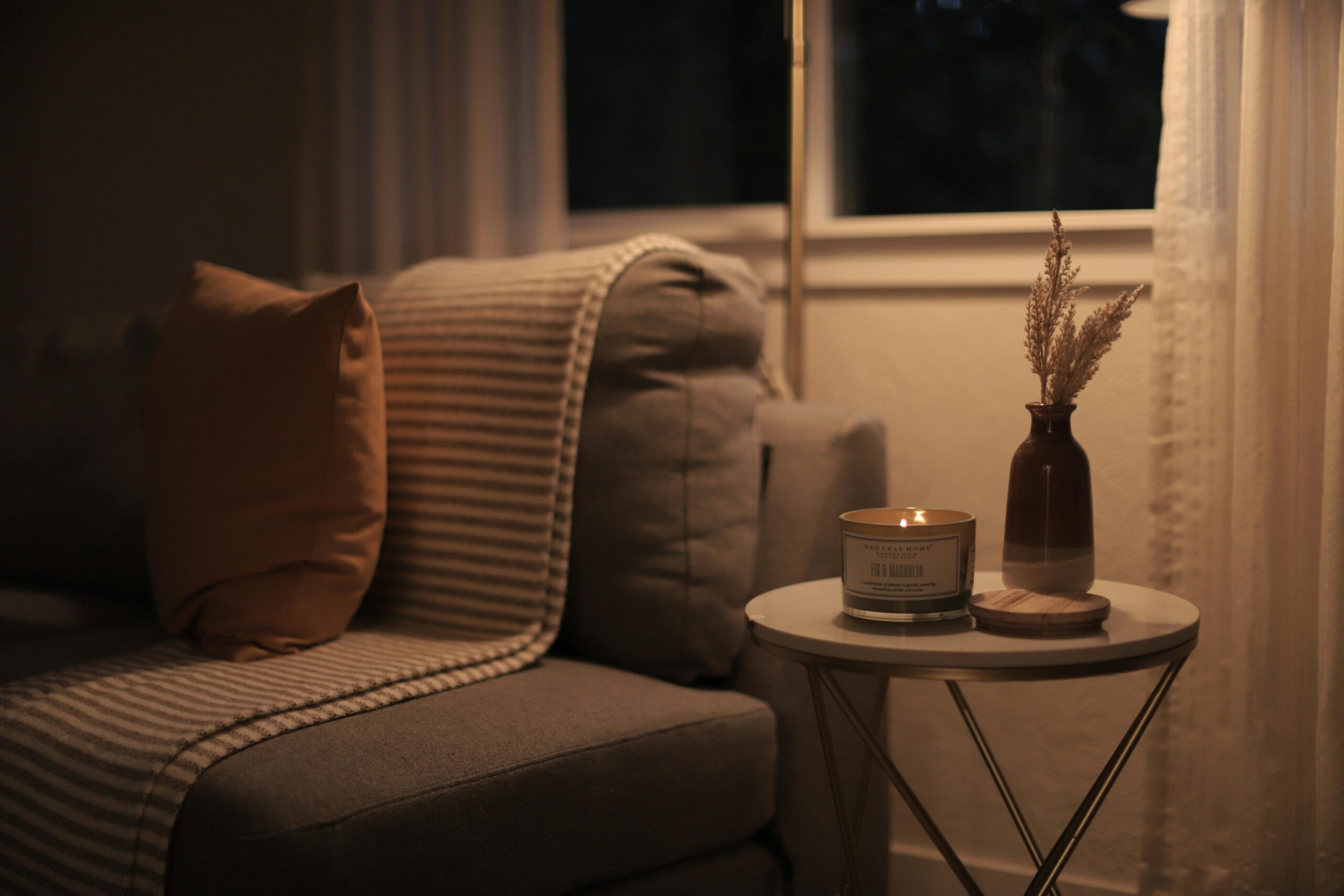 Przytulny kącik do czytania w stylu hygge z beżowym fotelem, ciepłym oświetleniem świecy na okrągłym stoliku, suszonymi trawami w ciemnym wazonie przy oknie wieczorem